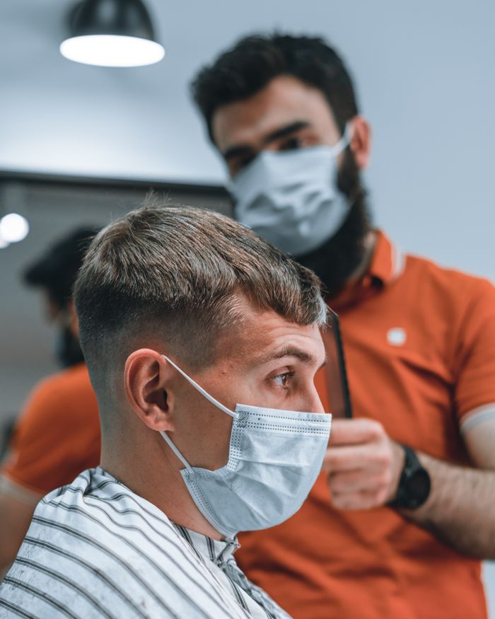 social-feed-img-05 Side view of a barber cutting hair of a client wearing a face mask in a modern barbershop.