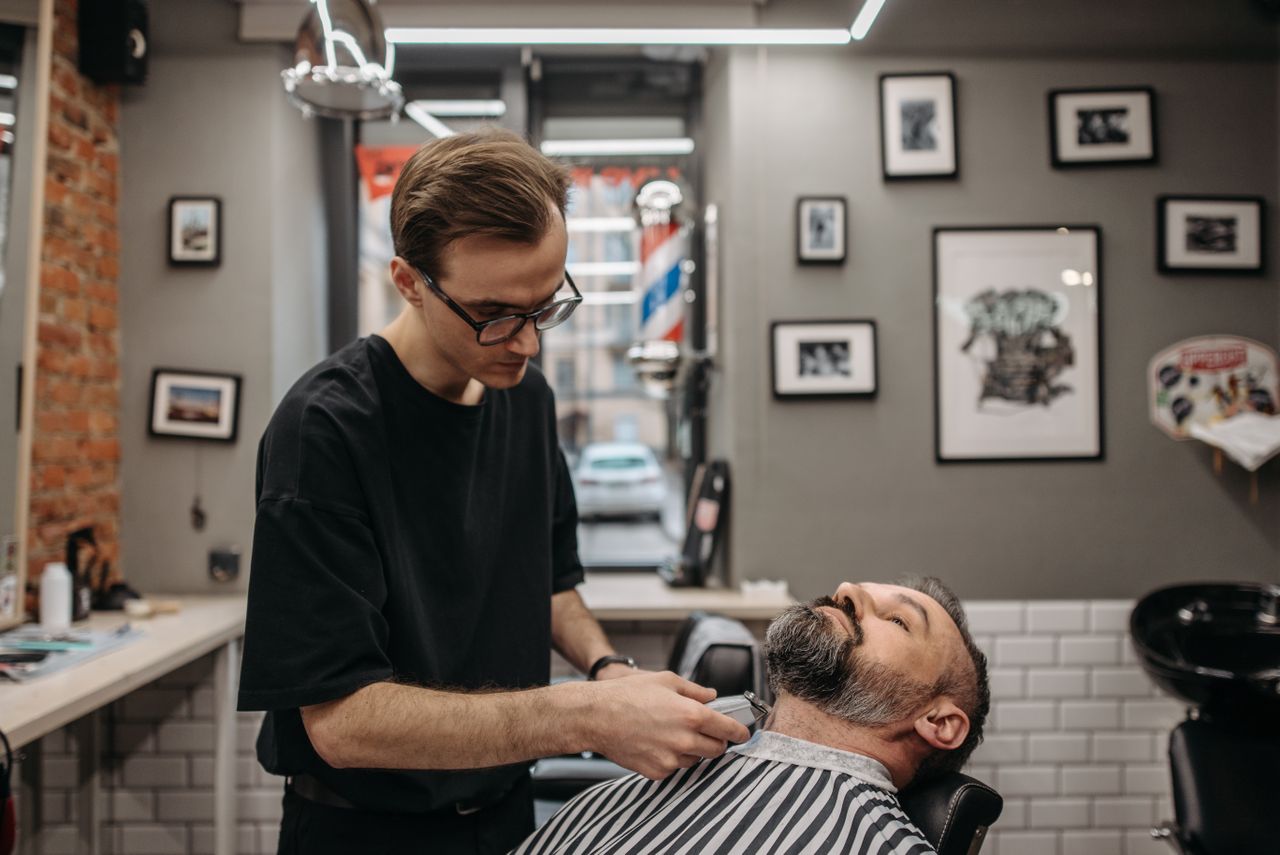 social-feed-img-03 A barber meticulously trims a bearded man's hair in a contemporary barbershop setting.